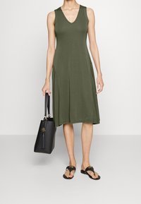 Kobieta prezentuje oliwkową sukienkę bez rękawów, trzymając czarną torebkę i nosząc czarne klapki, na neutralnym tle.