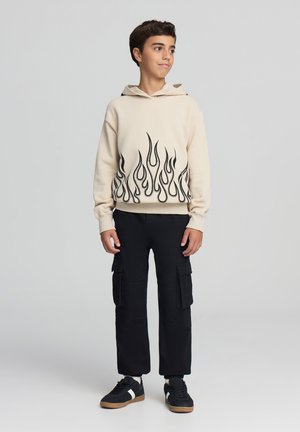 Felpa beige con grafica di fiamme nere sul bordo inferiore, abbinata a pantaloni cargo neri e sneakers nere con dettagli bianchi. Look casual.