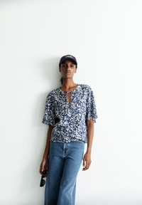 Marinebluse mit kurzen Ärmeln, Schlüsselhalsschnitt, strukturiertem Stoff, kombiniert mit hellblauen weiten Jeans und einer Sonnenbrille in der Hand.