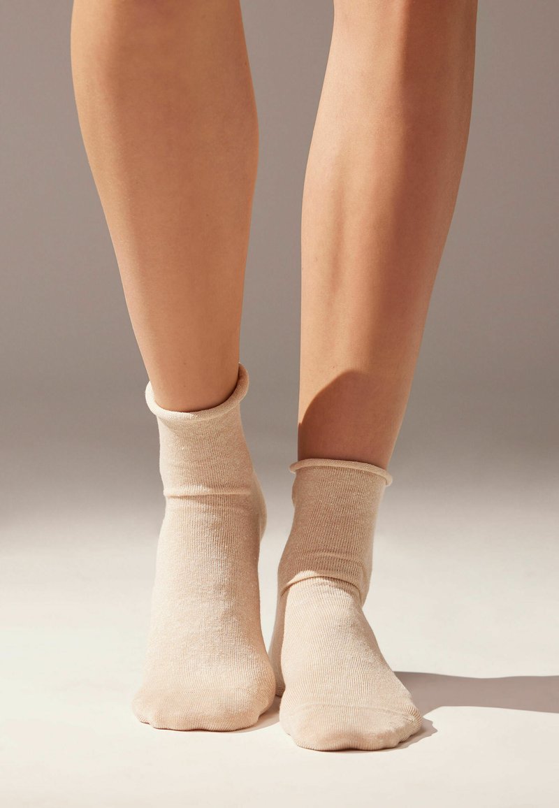Beige knöchellange Socken aus weichem, strukturiertem Stoff mit einem gerollten Bund, auf baren Füßen vor einem neutralen Hintergrund gezeigt.