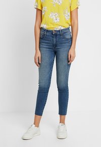 Jeans ajustados en denim azul oscuro, con cintura alta y pierna corta. Combinados con una camiseta de estampado floral amarillo brillante y zapatillas blancas.