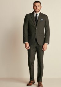 Traje entallado en verde oscuro con chaqueta de un solo boton, solapas tipo muesca y zapatos de cuero marrón. El pañuelo de bolsillo añade un contraste de color.