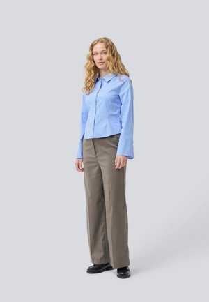 Chemise boutonnée rayée bleu clair à manches longues, associée à un pantalon large beige clair et des chaussures noires, se tenant devant un fond neutre.