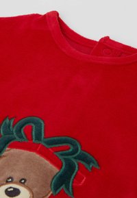 Vêtement en polaire doux rouge présentant un motif de visage d'ours avec des accents verts. Encolure ronde avec une fermeture à pression à l'arrière.