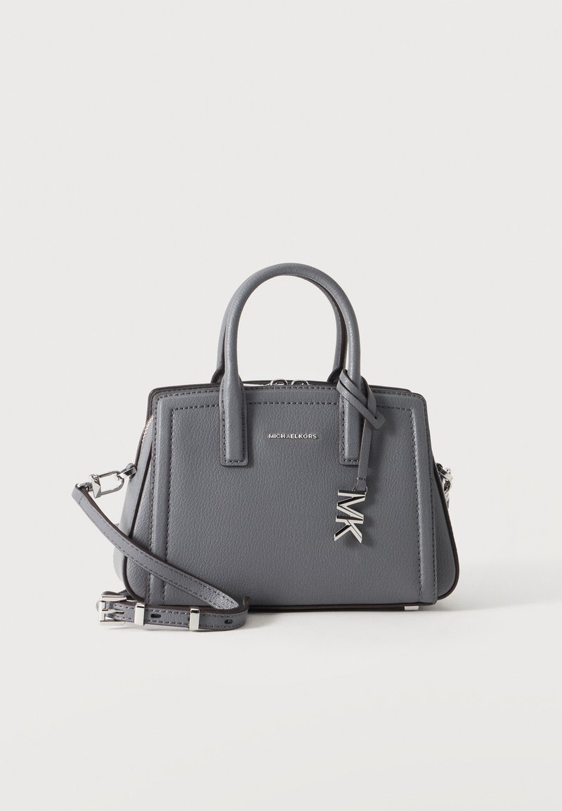 Γκρι τσάντα Michael Kors με πάνω χερούλια, αποσπώμενο ιμάντα ώμου και ασημί γούρι MK μπροστά, σε απλό λευκό φόντο.