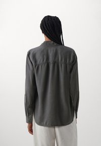 Mörkgrå skjorta med långa ärmar, knäppning vid ärmsluten och rundad nederkant. Har en ryggsöm och avslappnad passform. Slät textur.