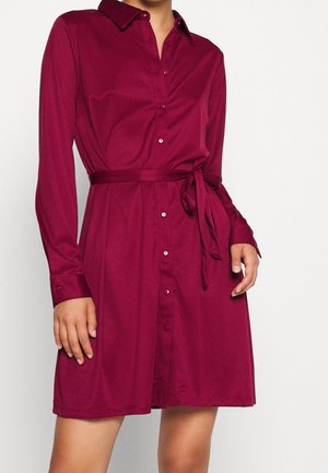 Femme portant une robe chemise à manches longues, boutonnée, rouge foncé, avec une ceinture nouée à la taille et un col classique.