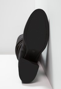 Svart mocka ankelsko med en texturerad gummisula, med en slouchad design och storleksmarkering 37 på botten.