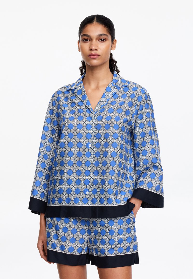 Femme portant un haut de pyjama bleu à motifs floraux à manches longues et un short assorti avec bordure noire, debout sur fond blanc.