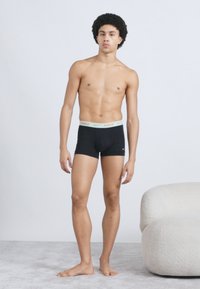 Mustad bokserid heleda oranži Nike logoga vööl, esitletud meeste modellil ühevärvilise tausta ees.