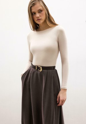Femme portant un haut à manches longues beige rentré dans une jupe marron taille haute avec une ceinture noire et une boucle dorée, debout avec une main dans la poche.