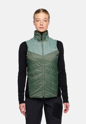Isolierte grüne Weste mit einem Zickzack-Steppdesign, hohem Kragen und Frontreißverschluss, kombiniert mit einem schwarzen Langarmshirt darunter.