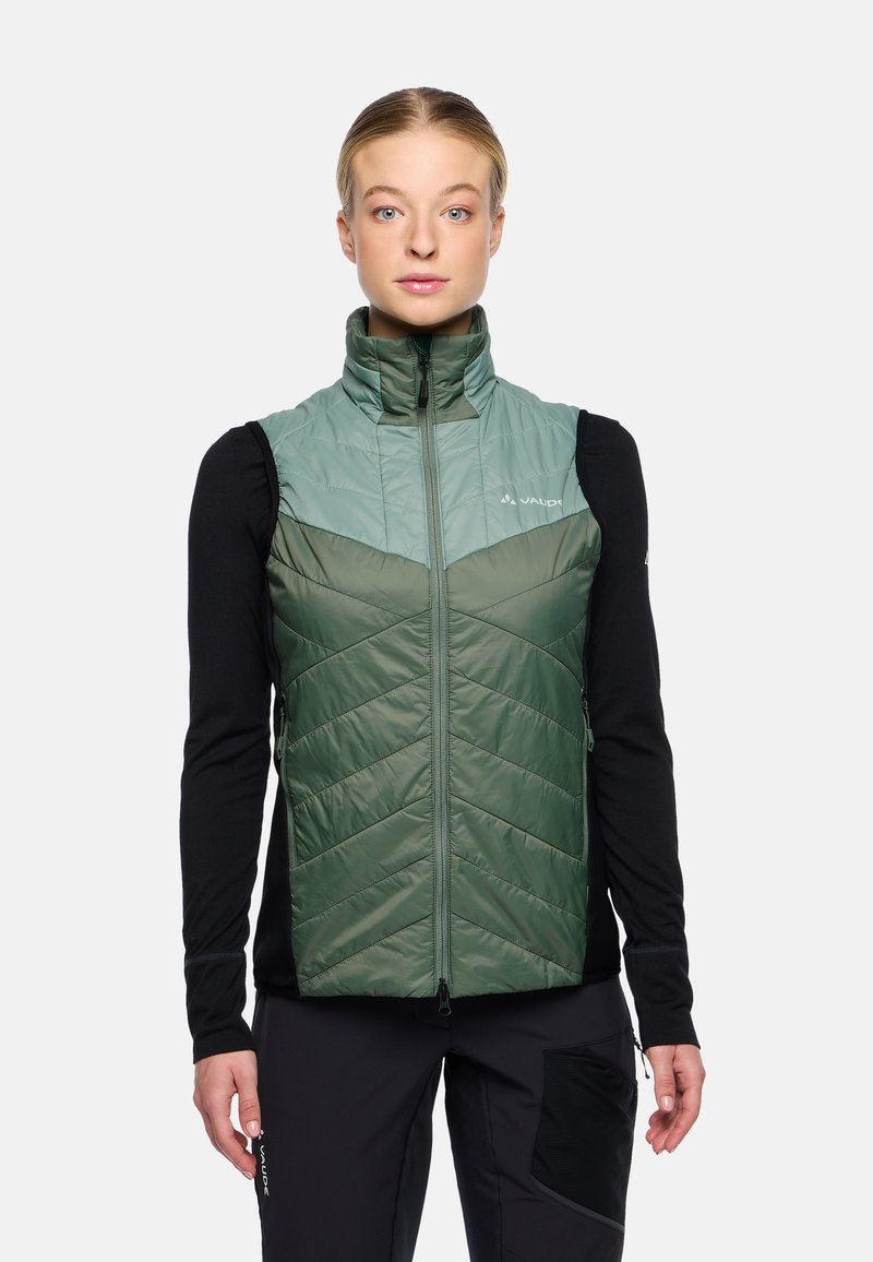 Gilet verde isolato con design a trapunto a zigzag, colletto alto e zip frontale, abbinato a una maglietta nera a maniche lunghe sotto.