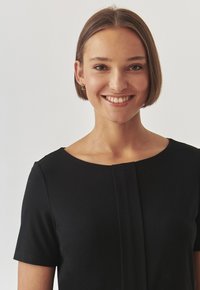 Must CSS-särgiv kaelusega must kleit lühikeste varrukatega ja voltidega esiküljel. Sile kangastekstuuri, hästi istuv korpus ja minimalistlik disain.