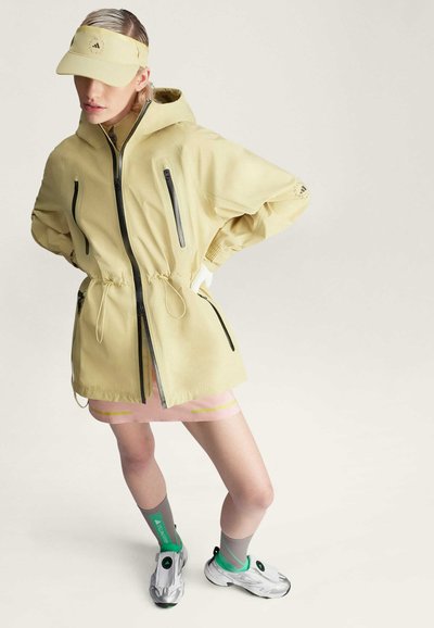 Chaqueta amarilla claro con capucha, bolsillos con cremallera y cintura ajustada. Combinada con una falda rosa, calcetines deportivos grises y calzado con acentos verdes.