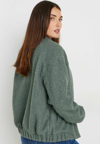 Green, fluffy fleece jacket with a relaxed fit, ribbed hem, and dropped shoulders. Textured surface and simple design, suitable for casual wear.