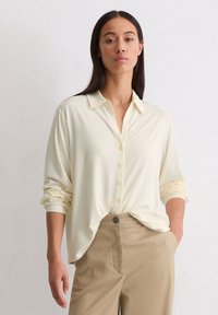 Vrouw met lang, donker haar die een crèmekleurig overhemd met opgerolde mouwen en beige broek draagt, tegen een effen lichte muur.