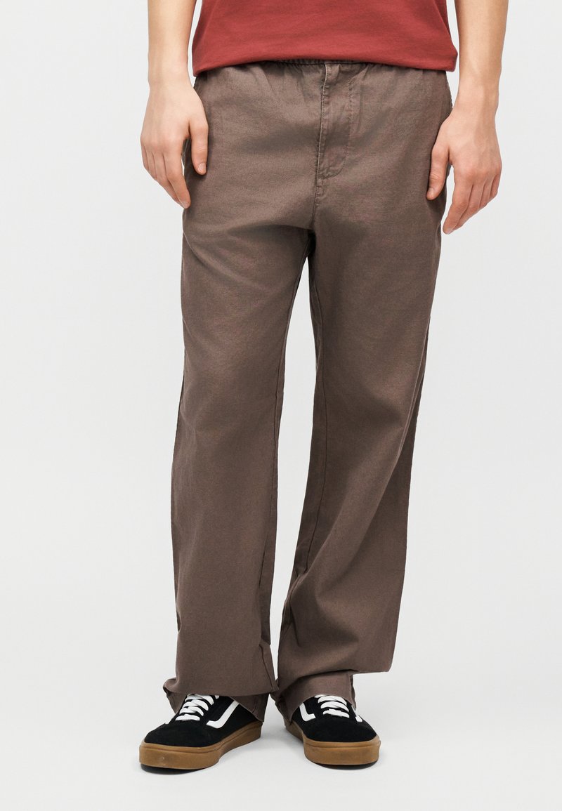 Άτομο που φοράει χαλαρά καφέ παντελόνια, μαύρα και άσπρα αθλητικά παπούτσια και κόκκινο πουκάμισο· στέκεται μπροστά σε απλό ανοιχτό φόντο.