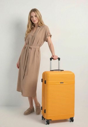 Młoda kobieta w beżowej sukience stojąca obok żółtego walizki na kółkach z wysuniętą rączką na jednolitym tle.