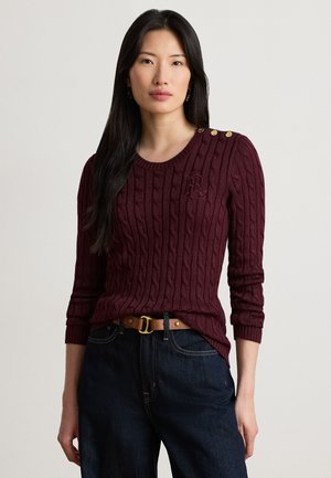 Femme aux longs cheveux noirs portant un pull torsadé bordeaux avec des boutons dorés et un jean bleu foncé sur un fond clair uni.