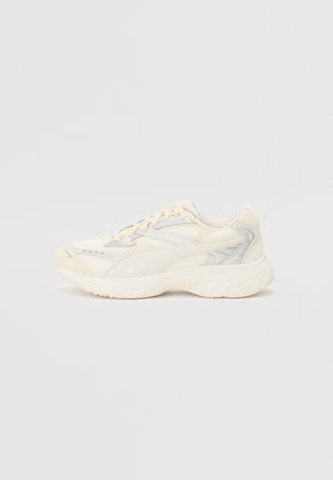 Chaussures de sport beige avec une tige en mesh et synthétique, une semelle incurvée et des accents réfléchissants. Elles présentent un design à lacets et une languette de traction à l'arrière.