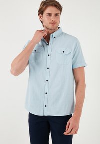 Camisa de botón de manga corta azul claro con dos bolsillos frontales, botones negros y un acabado de tejido texturizado. Cuello bien planchado.