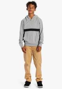 Grå huvtröja med en svart horisontell rand, dragkedja framtill och ribbade ärmslut, kombinerad med ljusbeiga byxor och svarta sneakers.