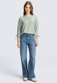 Svijetlozeleni pleteni top s teksturom, širokog kroja i vezicama na rukavima, uparен sa širokim plavim trapericama i svijetlim cipelama.