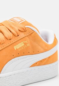 Orange suede sneaker featuring white accents, textured laces, and a gold logo. Design includes a rubber sole and visible stitching details.