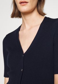 Marinblå kortärmad kofta med räfflad textur och V-ringning, med en knappknäppning framtill.