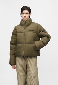 Person wearing an olive green puffer jacket with high collar and light beige cargo pants, standing against a plain white background.