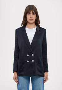 Blazer in satin nero con chiusura a doppio petto, rever a punte e due tasche frontali, abbinato a una camicia bianca e jeans blu.