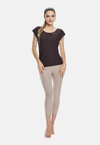 Melna īsām piedurknēm tops ar apaļu kakla izgriezumu un zelta logo; ķermeņa krāsas pieguļošas capri Legging bikses; modelis stāv basām kājām uz balta fona.