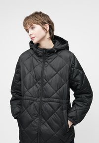 Veste noire rembourrée avec capuche, dotée d'un design matelassé en diamant, fermeture éclair, poignets élastiques et poches latérales. Texture du matériau lisse.