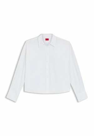 Camicetta bianca cropped con maniche lunghe e colletto classico. Caratterizzata da un'etichetta rossa con il marchio sul colletto e chiusure con bottoni.