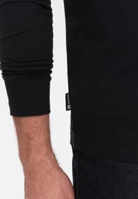 Maglietta nera a maniche lunghe con polsini aderenti, in morbido cotone, con un piccolo etichetta del marchio sul lato vicino all'orlo.