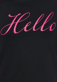 Camiseta negra con un gran texto en cursiva rosa que dice "Hola". La tela parece suave y tiene un diseño de cuello redondo estándar.