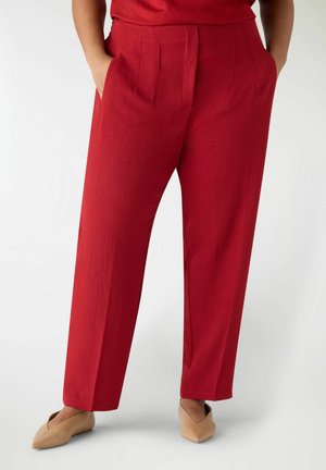 Donna che indossa pantaloni rossi a vita alta, gamba dritta, con tasche laterali, abbinati a scarpe piatte beige con punta appuntita, su uno sfondo neutro.