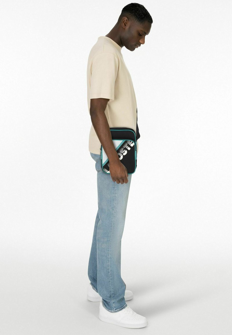 Sac rectangulaire noir et bleu sarcelle avec un motif graphique, porté sous le bras par une personne vêtue d'une chemise beige et d'un jean bleu clair.