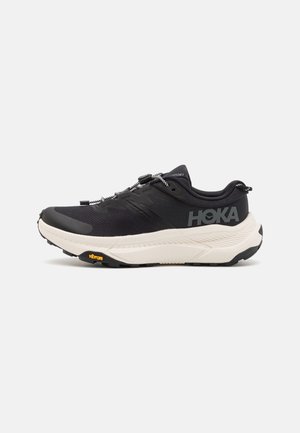 Chaussure de course Hoka noire et blanche avec semelle amortie et semelle extérieure Vibram, vue de côté sur fond blanc.
