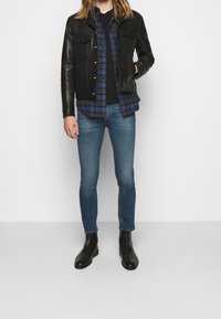 Svart läderjacka med tryckknappar, blå rutiga skjorta, svart T-shirt, blå slim-fit jeans och svarta ankelstövlar. Släta texturer och färger.