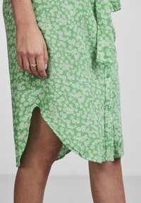 Grön klänning med vitt blommönster, knäppning fram och slits på sidan. Lättviktsmaterial med mjuk textur, knytning vid midjan.