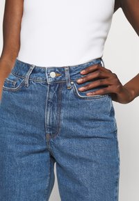 Vaqueros de mezclilla azul de tiro alto con un corte clásico, que cuentan con cierre de botón y cremallera, bolsillos delanteros y detalles de costuras en naranja.