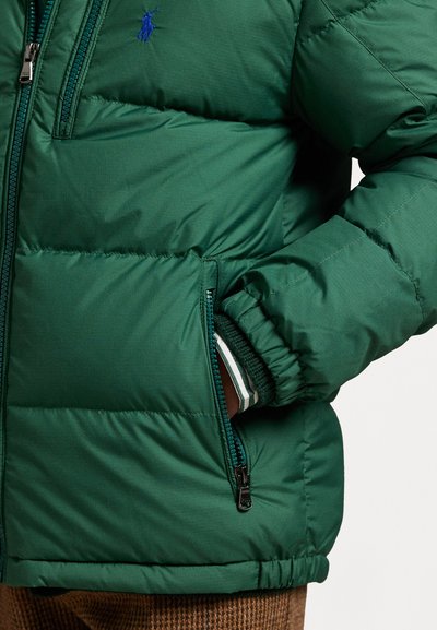 Grønn puffjakke med quiltet tekstur, som har en glidelåslomme, ribbestrikkede mansjetter med stripete aksent, og en logo-brodering på brystet.