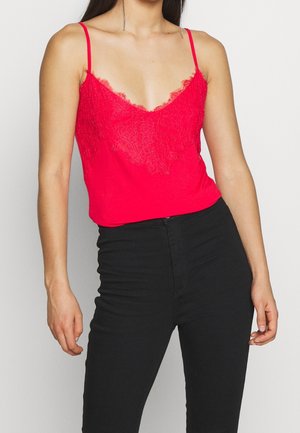 Rote Spitze-besetzte Camisole mit tiefem V-Ausschnitt und dünnen Spaghettiträgern, kombiniert mit engen schwarzen Hosen. Glatte Textur mit Spitzendetails.