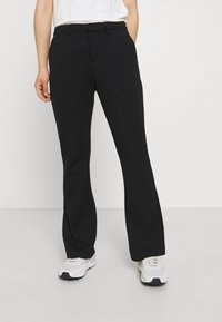Persona que lleva pantalones rectos negros y zapatillas blancas, de pie contra un fondo claro y liso.