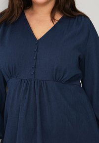 Navy blue long-sleeve dress with a V-neck, textured fabric, button detail at the neckline, and gathered waistline.