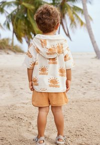 Brune fleece shorts og en kortærmet, cremet hættetrøje med et soltryk i orange, set fra ryggen mod en sandstrand.