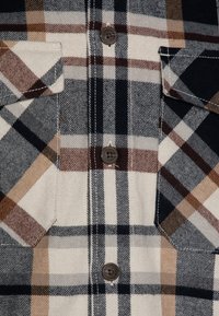 Chemise flanelle à carreaux présentant des tons de noir, blanc et marron. Conçue avec une patte de boutonnage et deux poches avant. Tissu texturé.