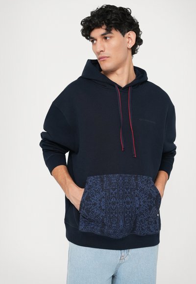 Marineblaues Hoodie mit einer gemusterten blauen Tasche, roten Kordeln und einer Kängurutasche. Weicher Stoff und lockere Passform, mit bedrucktem Logo.